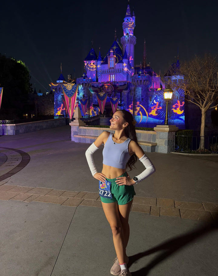 A woman posing in front of a brightly lit fantasy castle at night, wearing a light blue crop top, green athletic shorts with a race bib, and white arm warmers.