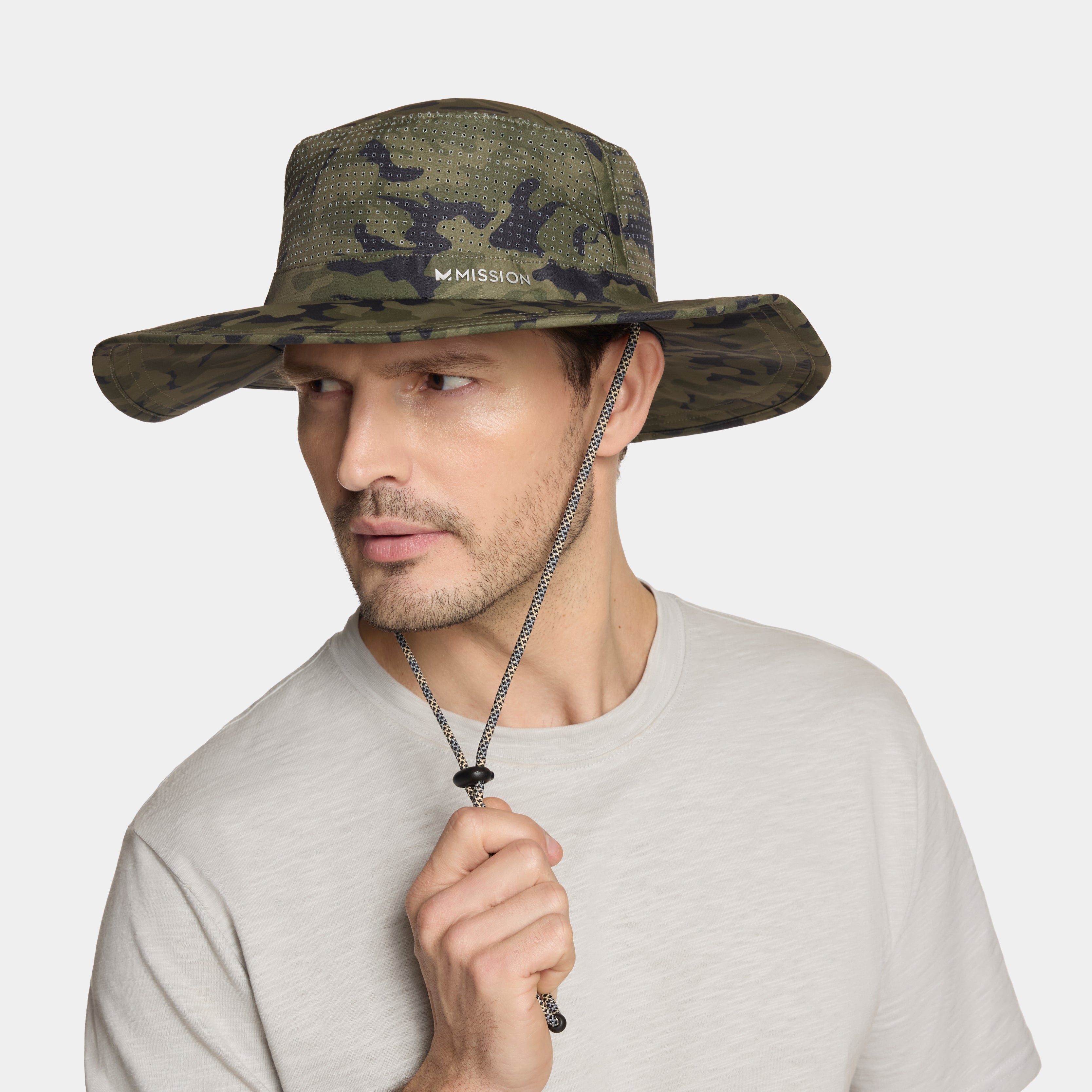 A man wearing a Mission brand camouflage boonie hat with a chin strap, paired with a light gray t-shirt, against a solid light gray background.