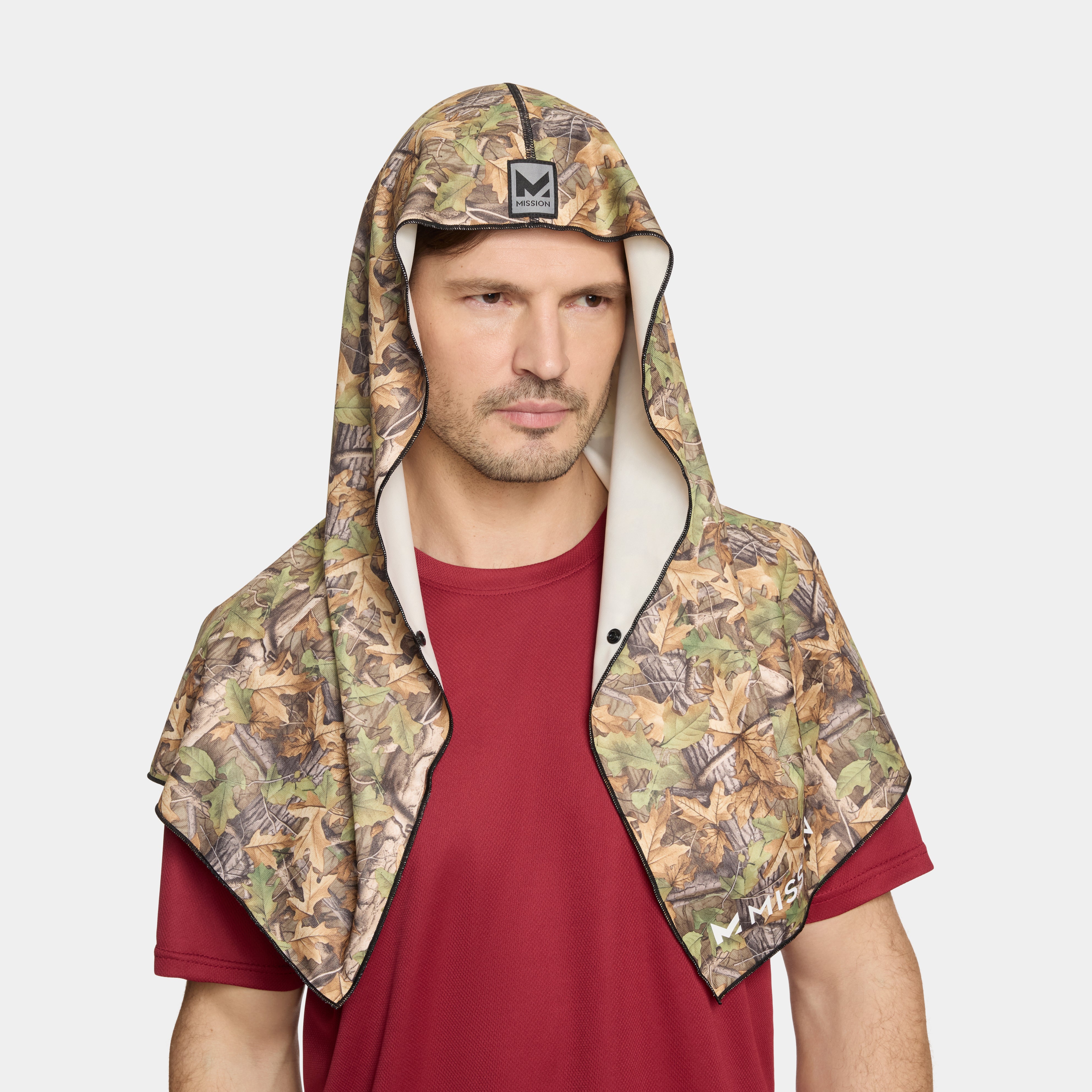 A man wearing a camouflage hooded neck warmer with a beige interior lining over a red t-shirt, shown against a light gray background.