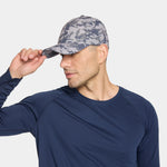 A man wearing a gray and blue camouflage baseball cap and a navy blue long-sleeve shirt, adjusting the brim of the cap against a light gray background.