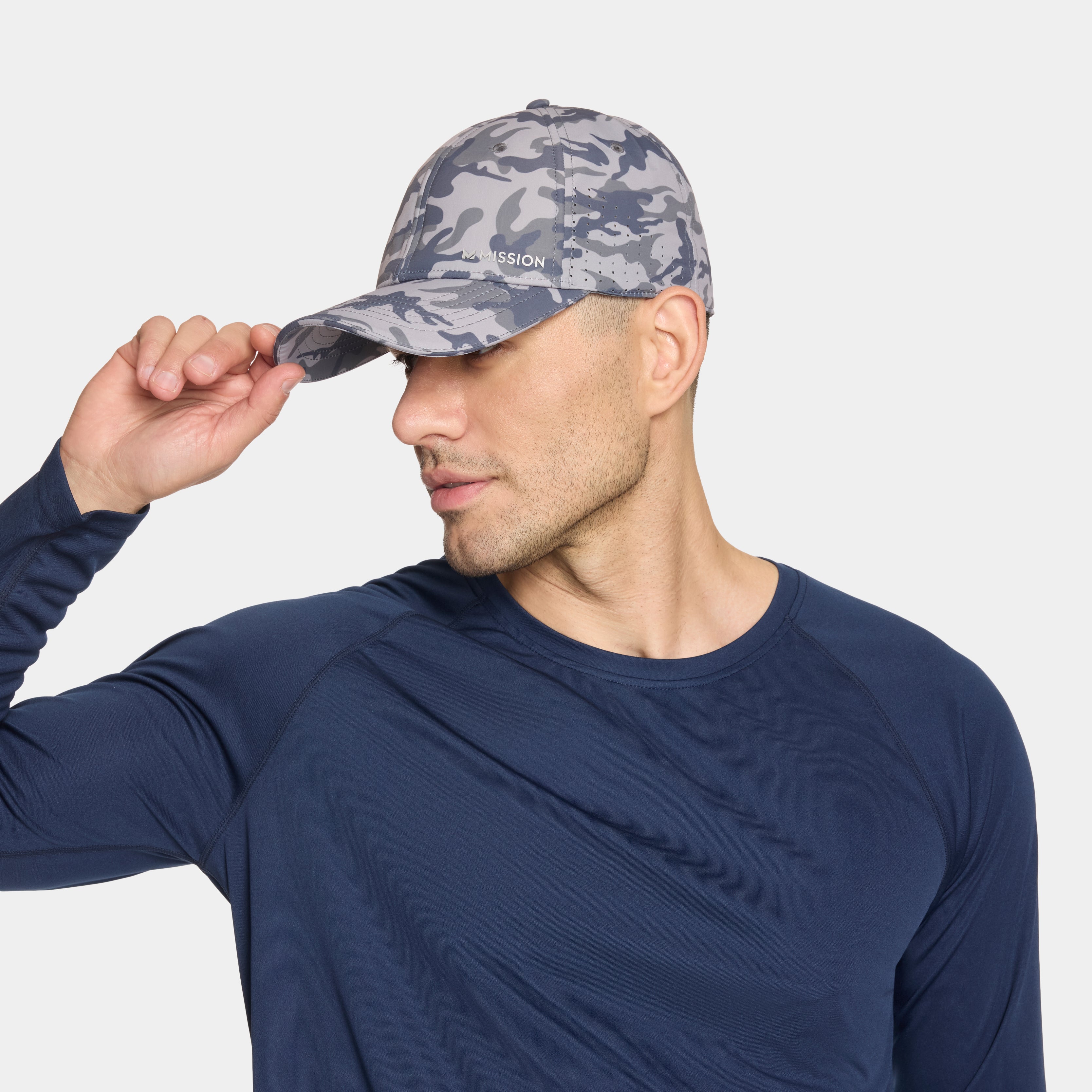 A man wearing a gray and blue camouflage baseball cap and a navy blue long-sleeve shirt, adjusting the brim of the cap against a light gray background.