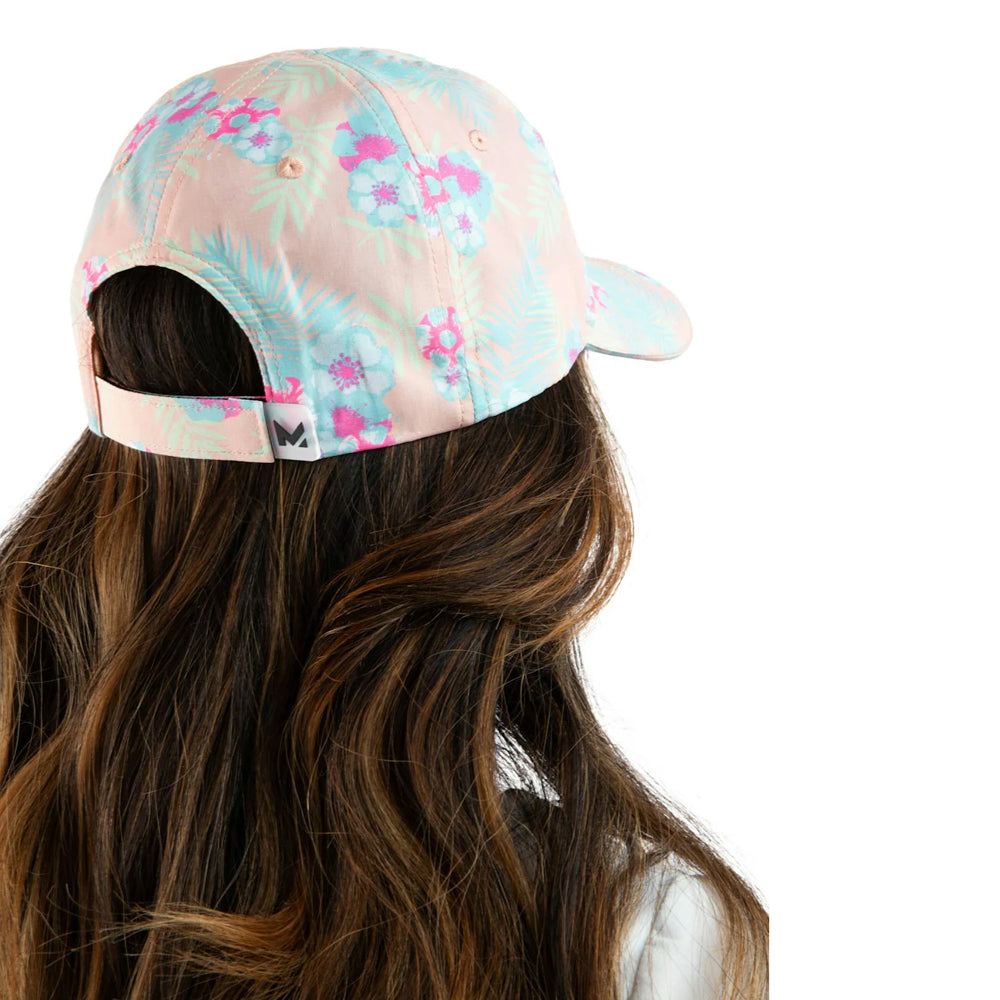 A woman shown from the back wearing a pastel pink baseball cap with a light blue and pink tropical floral print. The image is cropped to show her long brown hair and the back of the cap against a solid white background.