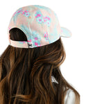 A woman shown from the back wearing a pastel pink baseball cap with a light blue and pink tropical floral print. The image is cropped to show her long brown hair and the back of the cap against a solid white background.