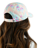 A woman shown from the back wearing a pastel pink baseball cap with a light blue and pink tropical floral print. The image is cropped to show her long brown hair and the back of the cap against a solid white background.