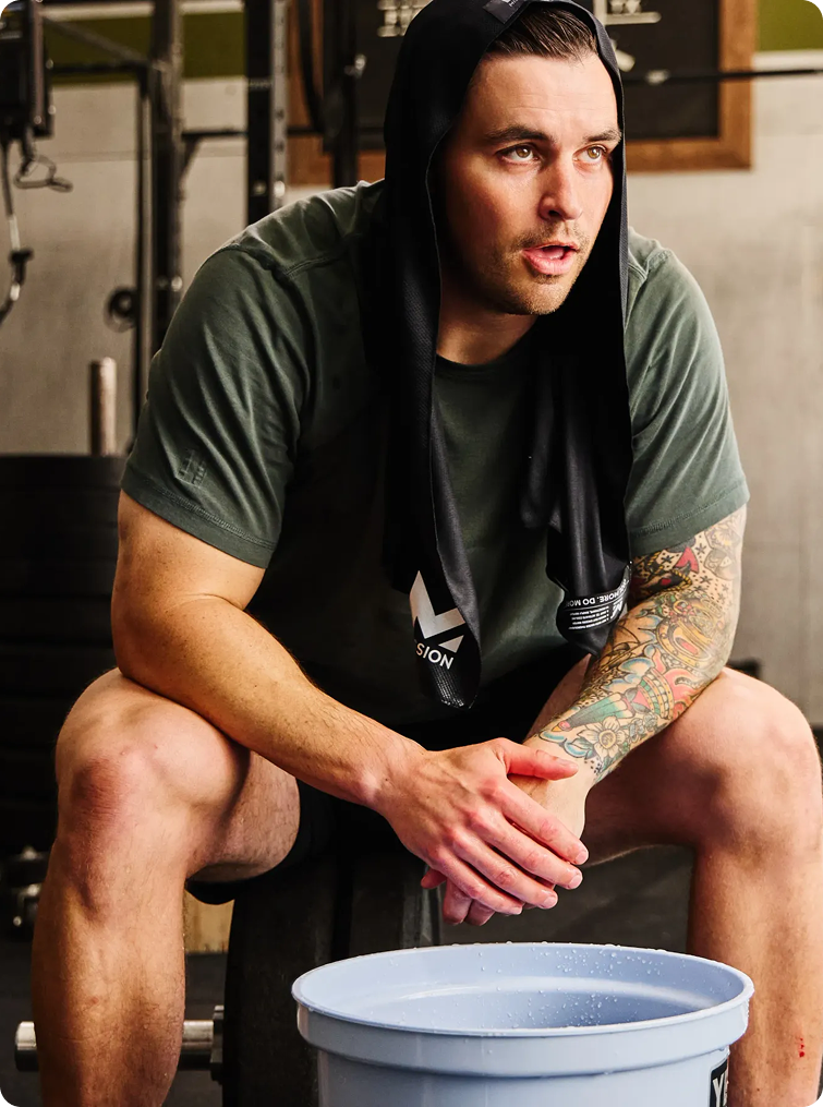 A man in a gym wearing an olive green t-shirt and a black hoodie draped over his shoulders, sitting on a weight bench with a blue bucket in the foreground.