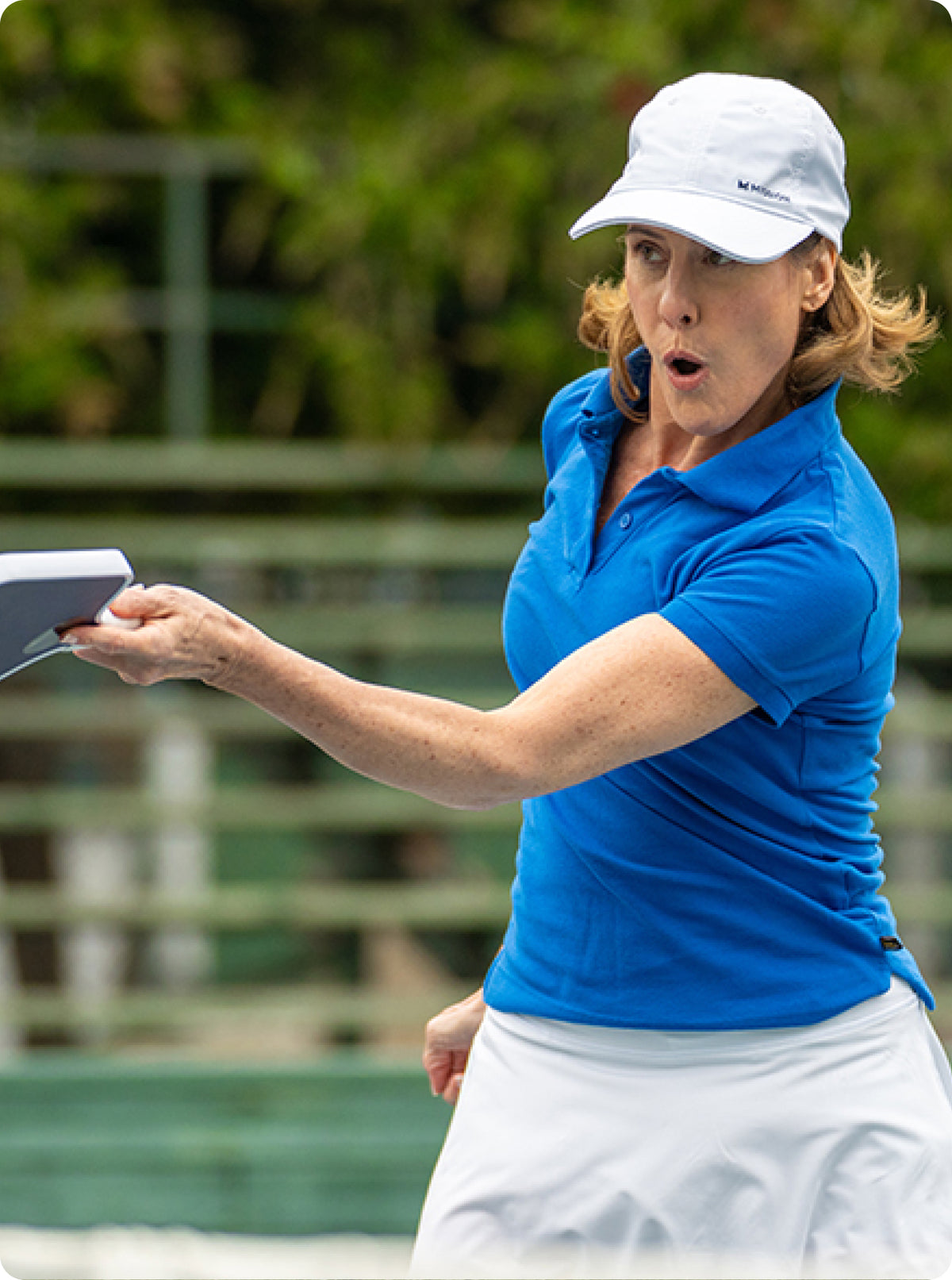 A woman on a tennis court wearing a bright blue polo shirt, white skirt, and white cap, captured mid-swing with a tennis racket.