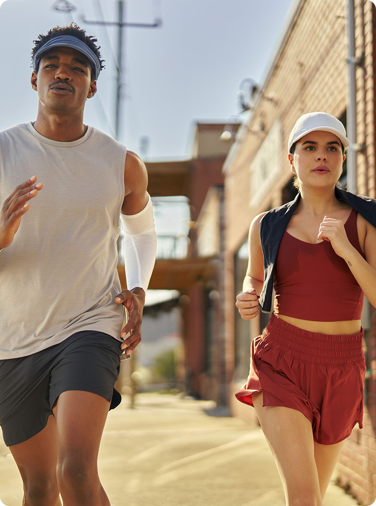 Two athletes jogging outdoors on a sunny urban street. The man wears a white sleeveless athletic top, dark gray running shorts, a blue visor, and a white arm sleeve.