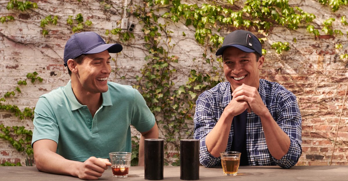 Two men laughing at an outdoor table, each wearing a dark navy baseball cap and a green or blue plaid polo shirt, with small glasses of tea in front of them against a brick wall with ivy.
