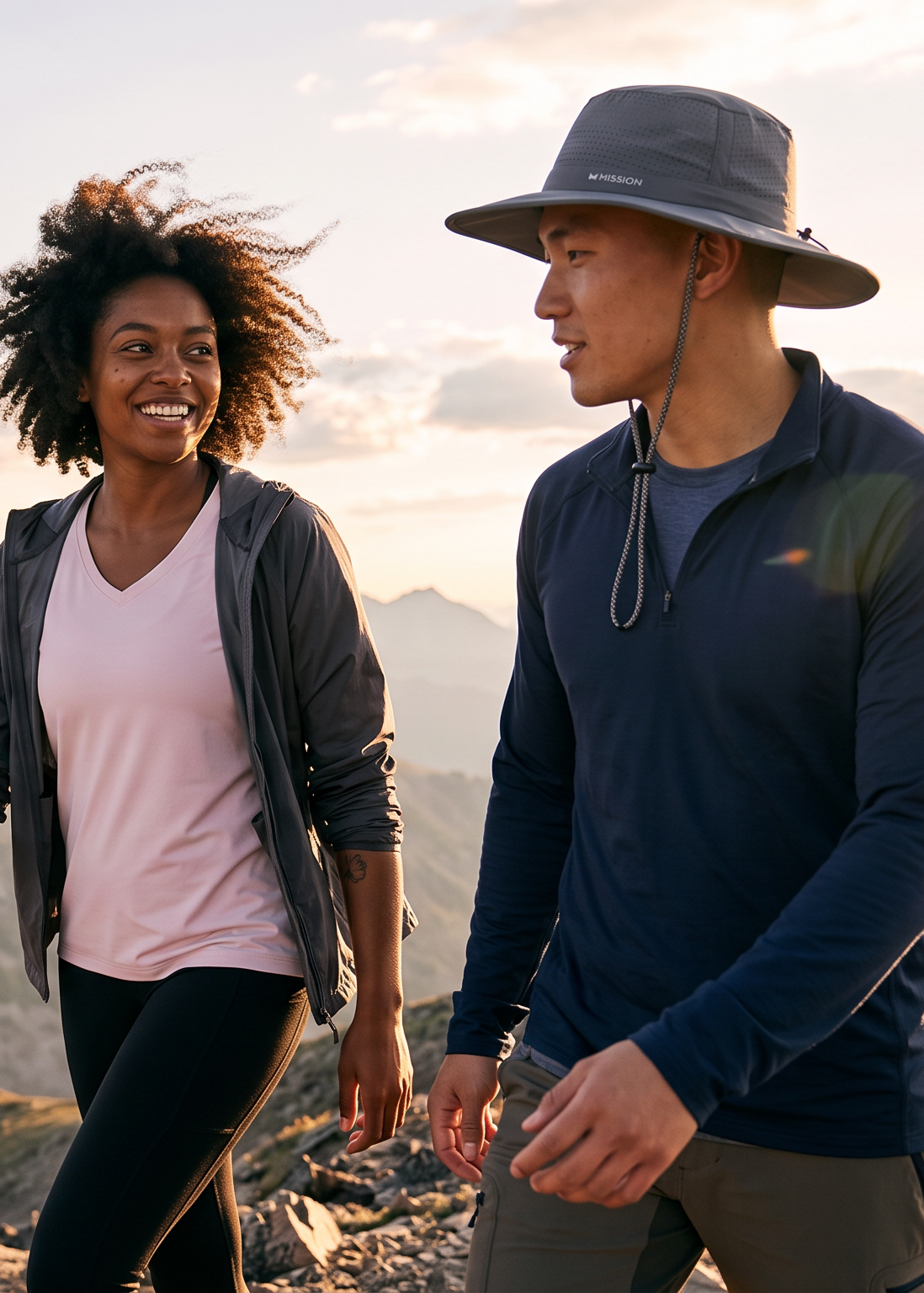 Two people hiking on a mountain ridge during golden hour, with rugged peaks and a cloudy sky in the background. One wears a wide-brim gray hat, navy quarter-zip top, and olive pants, while the other wears a light pink top, dark gray zip jacket, and black leggings.